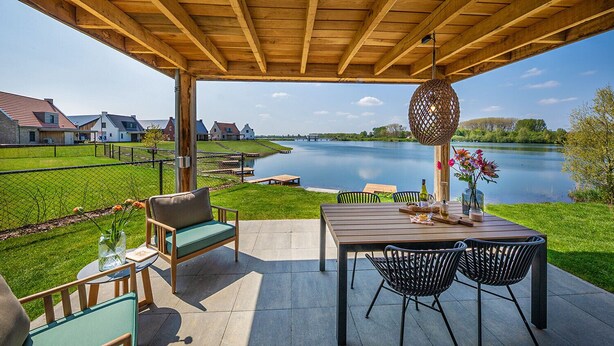 A covered patio with outdoor furniture, a table with a vase of flowers, and a view of a river and houses in the background. - Premium Water 4p - Two Bedroom Villa, Sleeps 4 (Thorn)