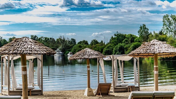 A beach with thatched umbrellas, lounge chairs, and a calm lake with boats in the background. - Luxe 6p - Three Bedroom Villa, Sleeps 6 (Thorn)