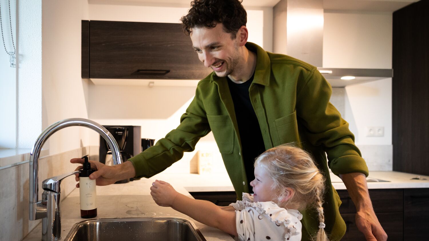 Une cuisine moderne avec un évier en acier inoxydable, une personne et un enfant interagissant près de l’évier.