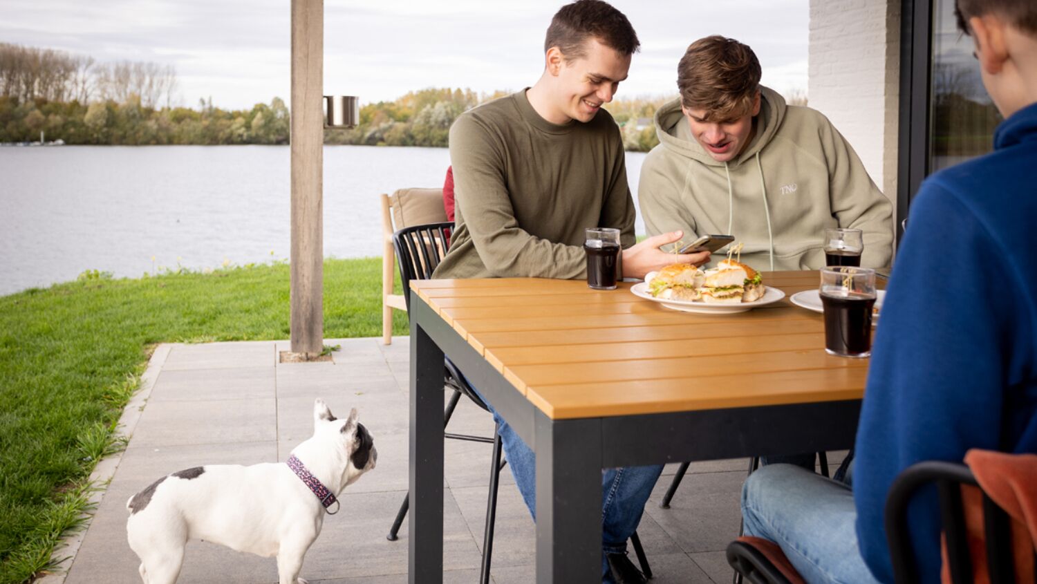 Una mesa con una hamburguesa y bebidas, un perro y vistas al lago.