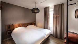 Een moderne hotelkamer met een houten hoofdeinde, een groot bed, een nachtkastje met een lamp, een hanglamp en uitzicht op het omliggende landschap door een raam met gordijnen.