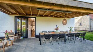 An outdoor patio with a wooden table and chairs, a glass door leading to an interior space, and a decorative wall hanging.