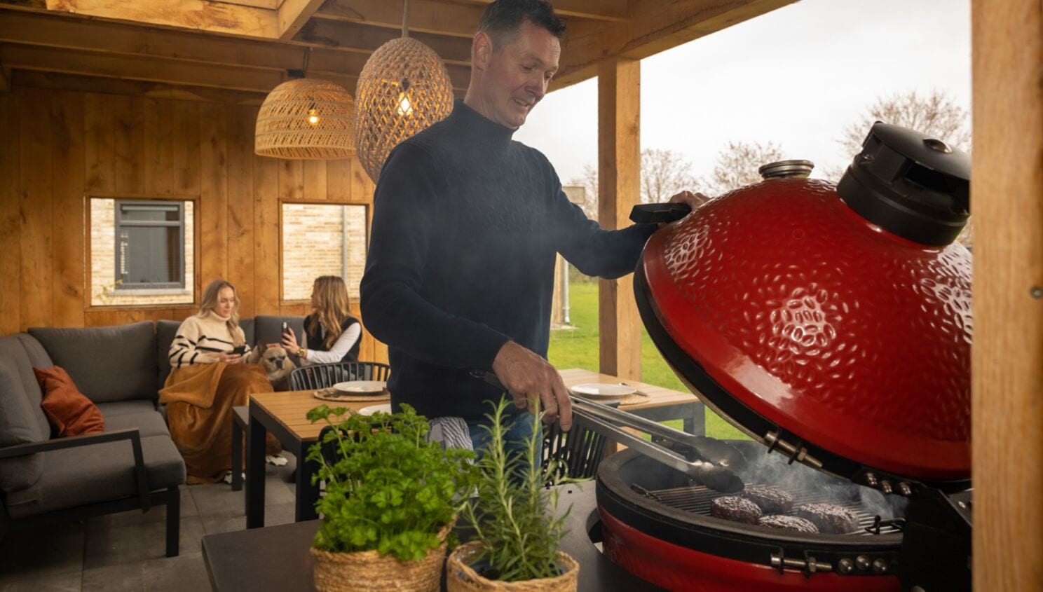 A person grilling outdoors with a red grill, a wooden interior, and a dining area with two people seated.
