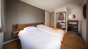 Una habitación de hotel con una cama grande, una mesita de noche con lámpara, un armario, un espejo y un baño con lavabo y espejo.