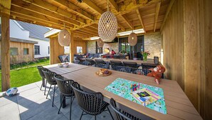 Un espacio de comedor al aire libre con mesa y sillas de madera, una lámpara colgante de fibras tejidas y vista a una casa al fondo.