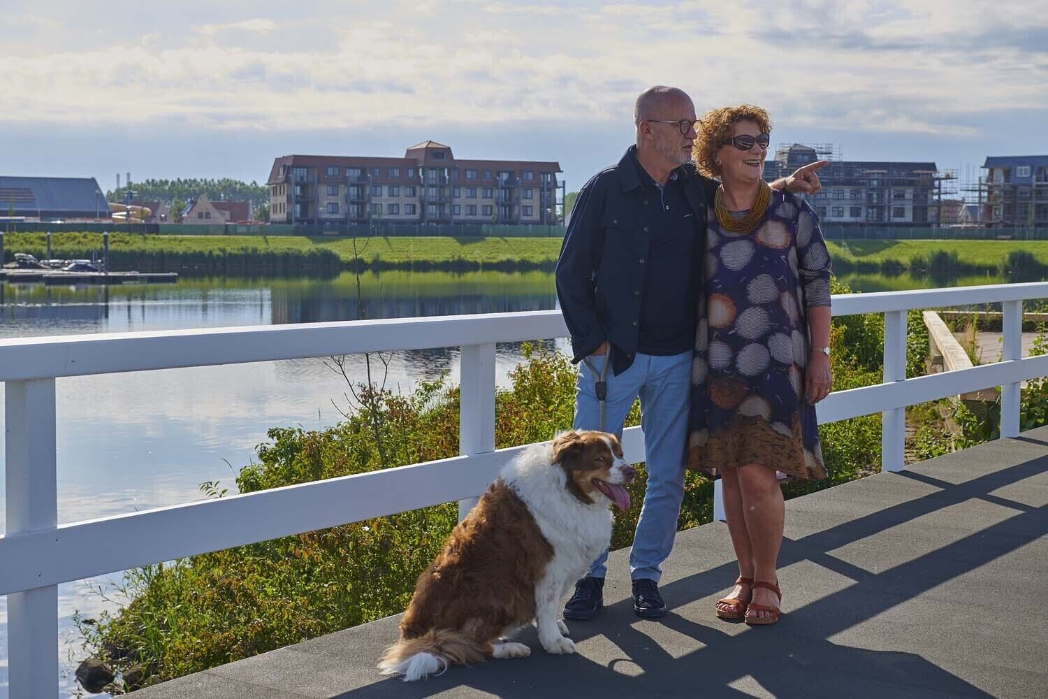 Ett par står på en bro med en hund, och i bakgrunden syns en vattenyta och några byggnader.