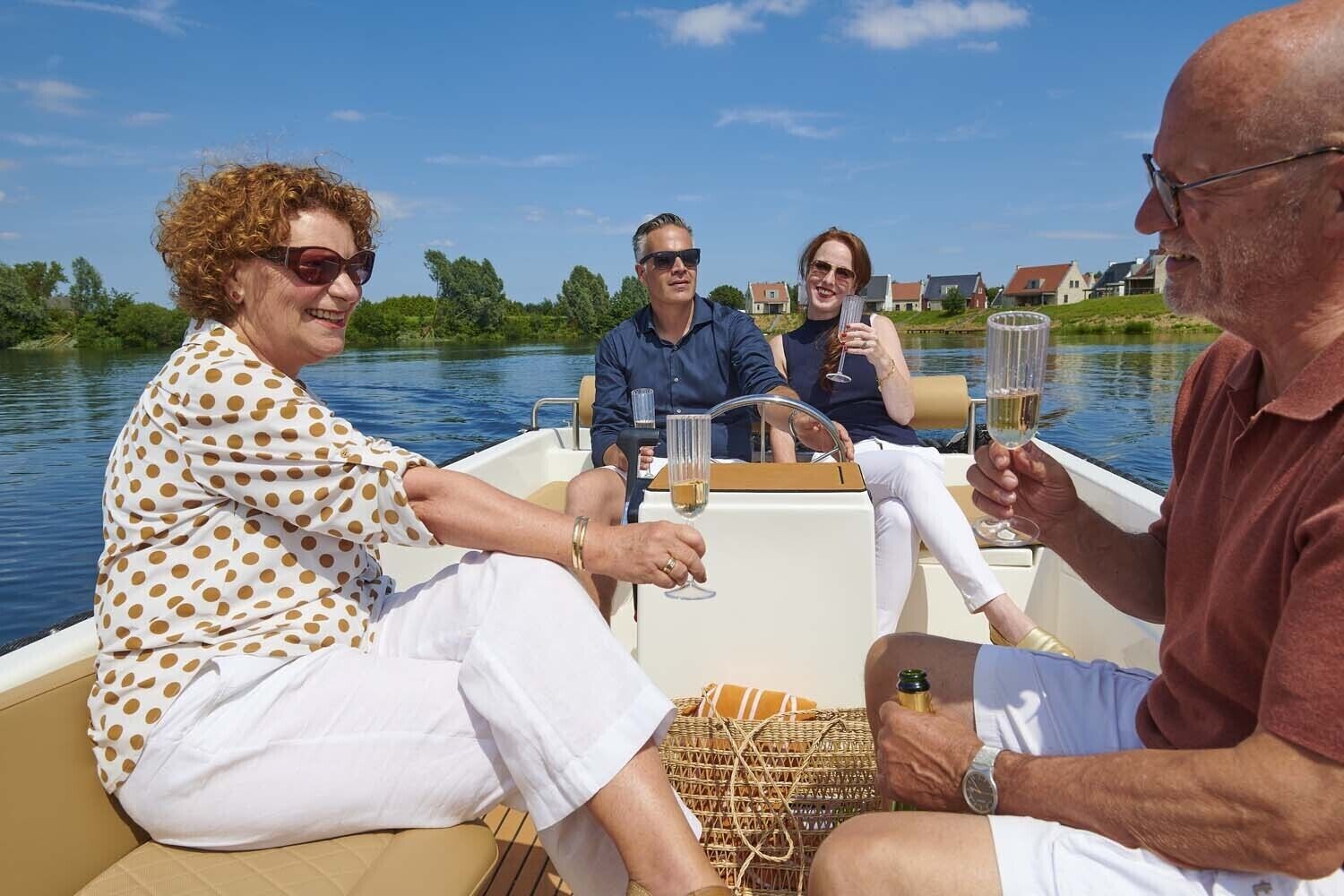 A boat with four people, each holding a glass of wine, on a calm body of water with houses in the background.
