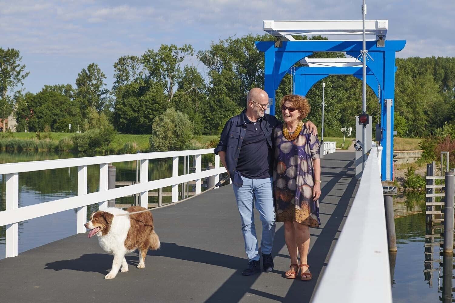 A couple walking on a bridge with a dog, surrounded by greenery and water.