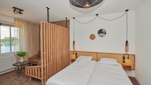 Moderni hotellihuone, jossa on suuri sänky, puinen väliseinä, pieni pöytä, jossa on kasvi, ja näkymä ulos.