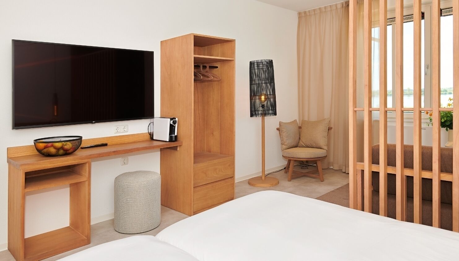 Camera d'albergo moderna con una grande TV a schermo piatto, arredamento in legno, un angolo salotto con una poltrona e vista separata da un pannello di legno.