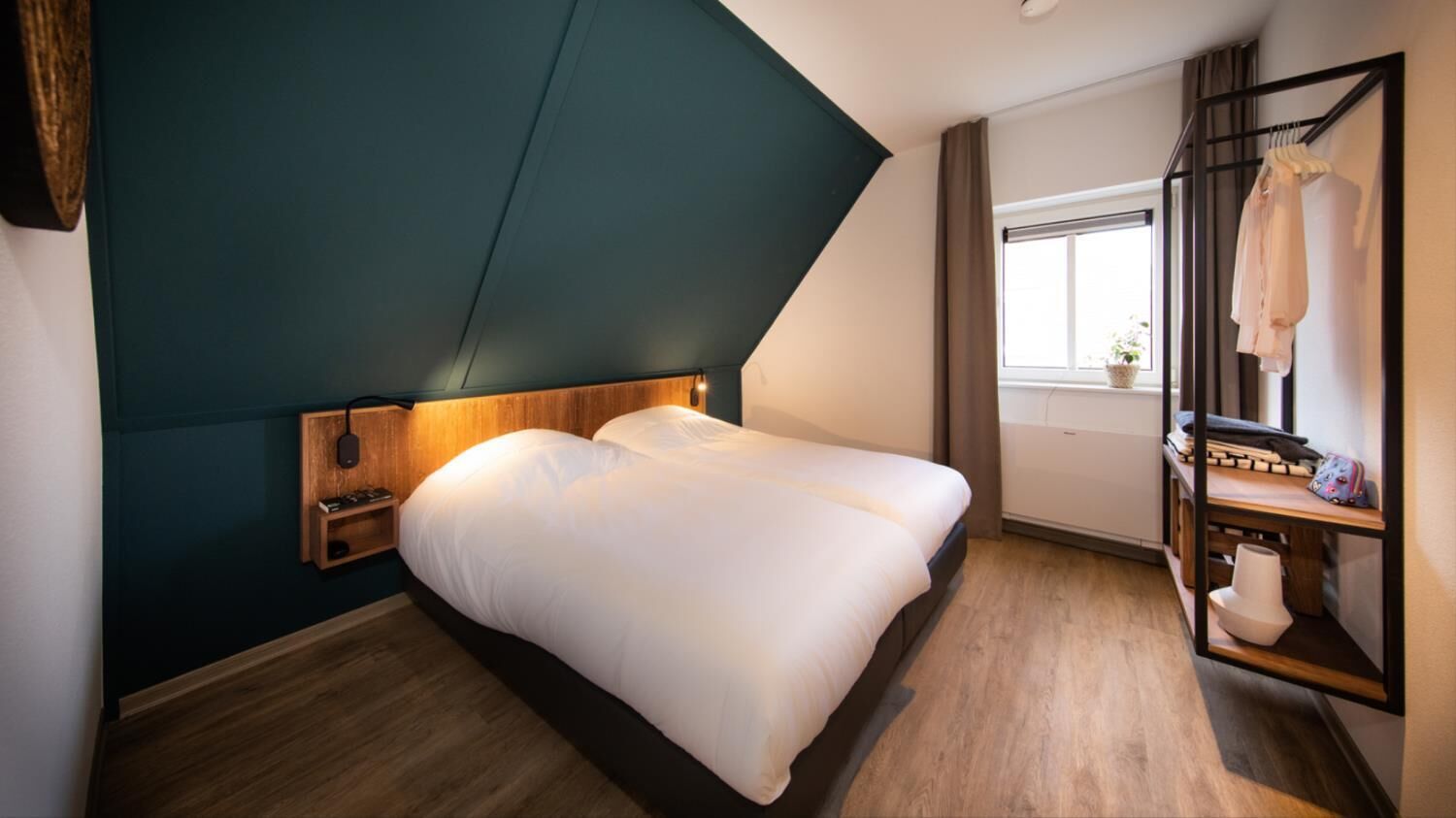 A modern bedroom with a bed, wooden headboard, bedside table, and a small shelf with a lamp.