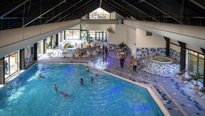 Une piscine intérieure dotée de plusieurs couloirs de nage, d’une zone dédiée aux plongeons et d’un espace détente avec des chaises et des tables.