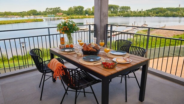 A dining table set on a balcony with a view of a lake, surrounded by chairs and a vase of flowers. - Lake View Maisonette 4p (Thorn)