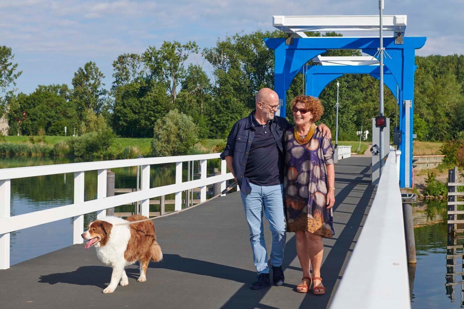 Uma ponte para pedestres com guarda-corpo branco, estrutura azul e um cão acompanhando duas pessoas.
