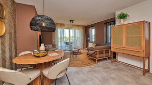 A modern living room with a wooden dining table, two sofas, a rattan cabinet, and a large window with curtains. - Lake View Maisonette 4p (Thorn)