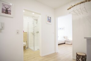 A modern hotel room with a wooden floor, a glass shower, a bed with a white headboard, and a small bedside table.