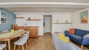Ein modernes Wohnzimmer mit einem blauen Sofa, einem Esstisch mit Stühlen und einer Küchenzeile mit Holzschränken und Regalen.