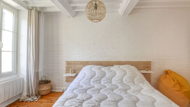 Ein Schlafzimmer mit weißer Ziegelwand, einem Holz-Kopfteil, weißer Bettdecke, einer gewobenen Pendelleuchte und einem Fenster mit Vorhängen.