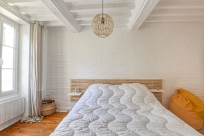 Un dormitorio con pared de ladrillo blanco, cabecera de madera, cubrecama blanco, lámpara colgante tejida y una ventana con cortinas.