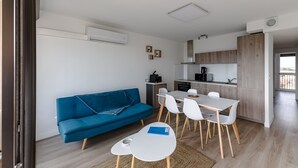 A modern living space with a blue sofa, white dining table, and chairs, a kitchenette, and a wooden floor. - Ombeline - Apt proche mer avec balcon (Saint-Cyprien)