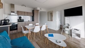 A modern kitchen with white cabinets, a dining area with a white table and chairs, a blue sofa, and a flat-screen TV mounted on the wall. - Ombeline - Apt proche mer avec balcon (Saint-Cyprien)