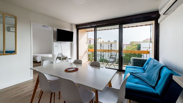 A modern room with a dining table, white chairs, a blue sofa, and a large window offering a view of buildings and a balcony. - Ombeline - Apt proche mer avec balcon (Saint-Cyprien)