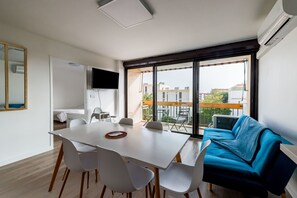 Een moderne kamer met een eettafel, witte stoelen, een blauwe bank en een groot raam met uitzicht op gebouwen en een balkon.