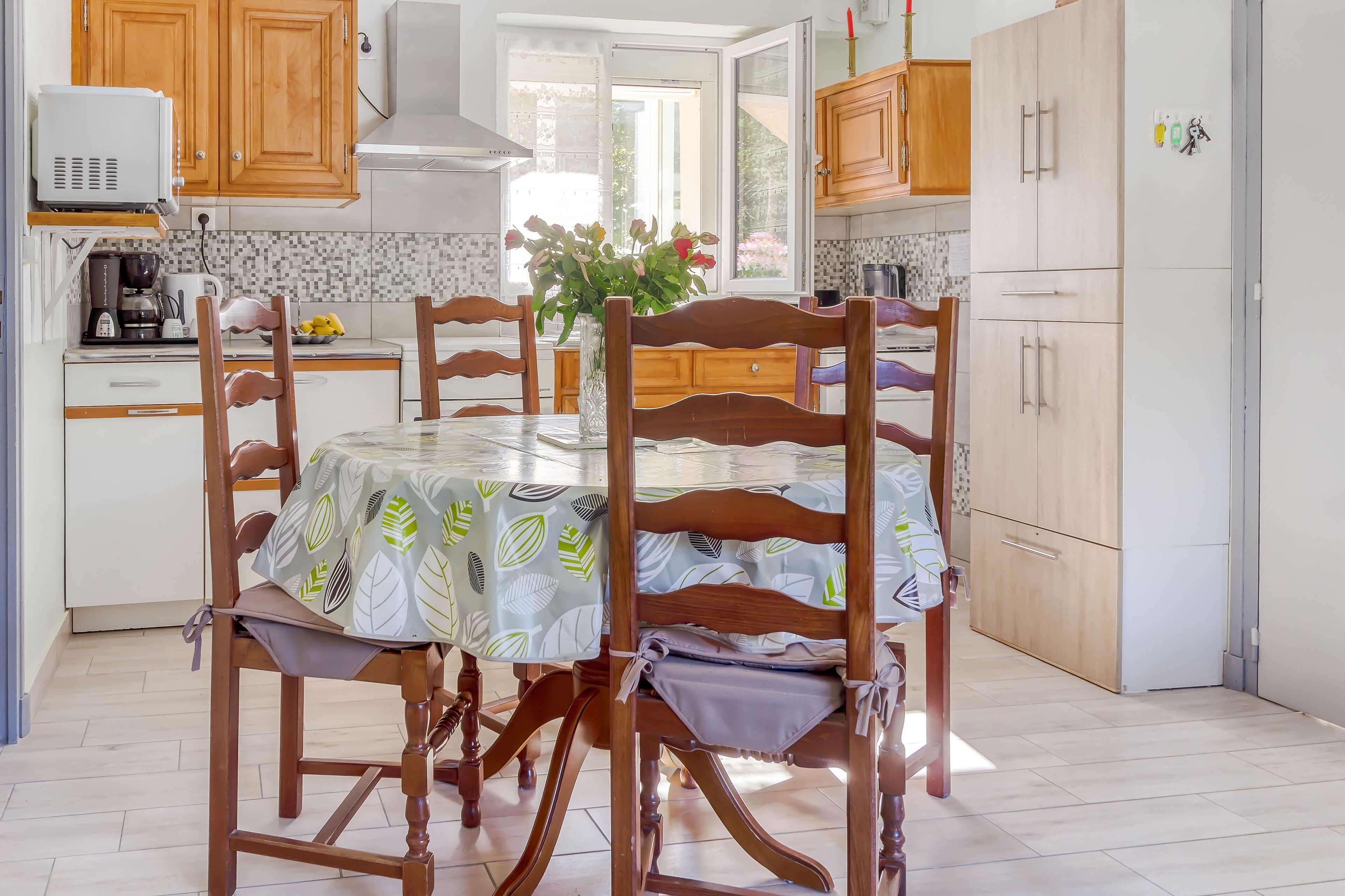 Une cuisine avec des meubles de rangement en bois, une table à manger recouverte d’une nappe à motifs de feuilles et un vase rempli de fleurs.