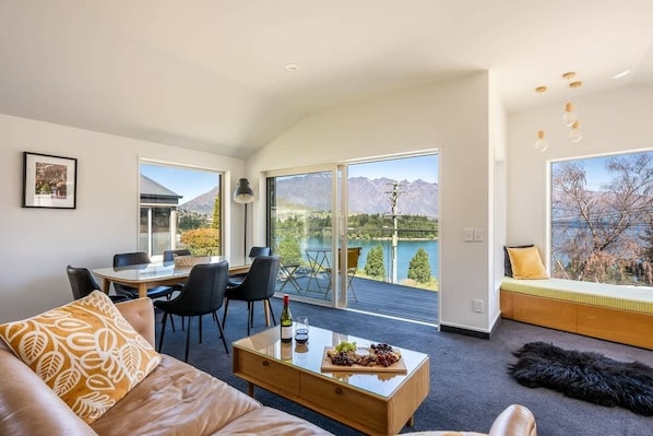 A modern living room with a sofa, coffee table, dining area, and a balcony with a view of mountains and a lake.