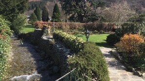 Een pad van stenen loopt door een tuin met een beekje, omringd door strakke hagen en bomen.