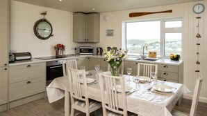 A kitchen with a dining area, a view of the countryside, and a clock on the wall. - Atlantis (Wadebridge)