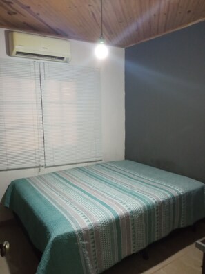 A room with a bed, an air conditioning unit, and a window with blinds.