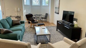 A modern living room with a green sofa, beige armchairs, a coffee table, a TV on a black stand, and a dining area with a table and chairs. - New 2 BR APT: amazing location, spotless, free private parking.  (Washington)