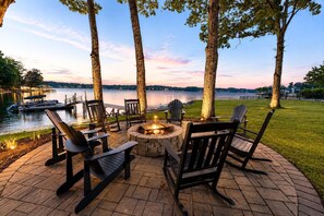 An outdoor seating area with Adirondack chairs, a fire pit, and a view of a lake with boats. - Channel Marker: Exclusive Lakefront Home w/Hot Tub, Custom Bunk Room, Dock & Optional Boat Rental (Mooresville)