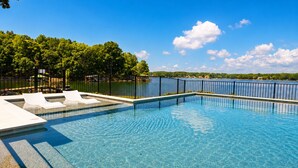 Une piscine d’un bleu éclatant, dotée de chaises longues, entourée d’une clôture et d’arbres, et offrant une vue sur un lac et des bâtiments situés au loin.