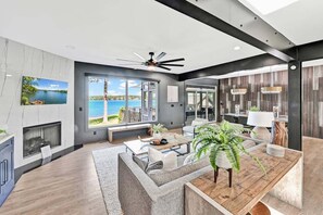 A modern living room with a large window overlooking a body of water, a fireplace, and a ceiling fan. - Channel Marker: Exclusive Lakefront Home w/Hot Tub, Custom Bunk Room, Dock & Optional Boat Rental (Mooresville)