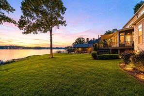 A waterfront property with a well-maintained lawn, a large tree, and a building with a balcony overlooking the water. - Channel Marker: Exclusive Lakefront Home w/Hot Tub, Custom Bunk Room, Dock & Optional Boat Rental (Mooresville)