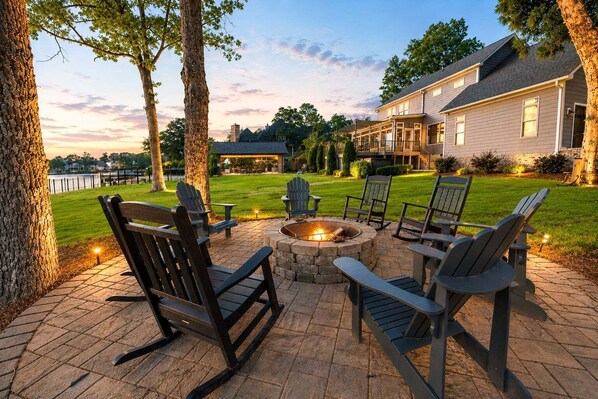 An outdoor seating area with Adirondack chairs and a fire pit, surrounded by a well-maintained lawn and a large house in the background. - Channel Marker: Exclusive Lakefront Home w/Hot Tub, Custom Bunk Room, Dock & Optional Boat Rental (Mooresville)