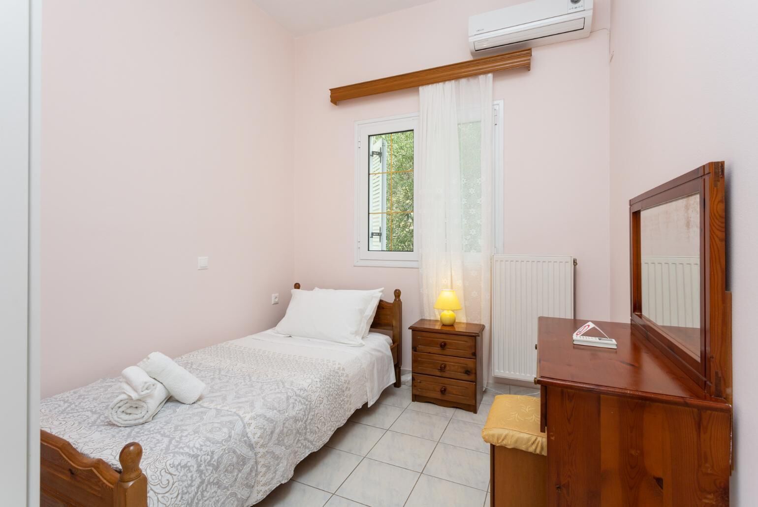 Une petite chambre à coucher avec un lit simple, une table de chevet en bois, une commode et une fenêtre avec des rideaux.