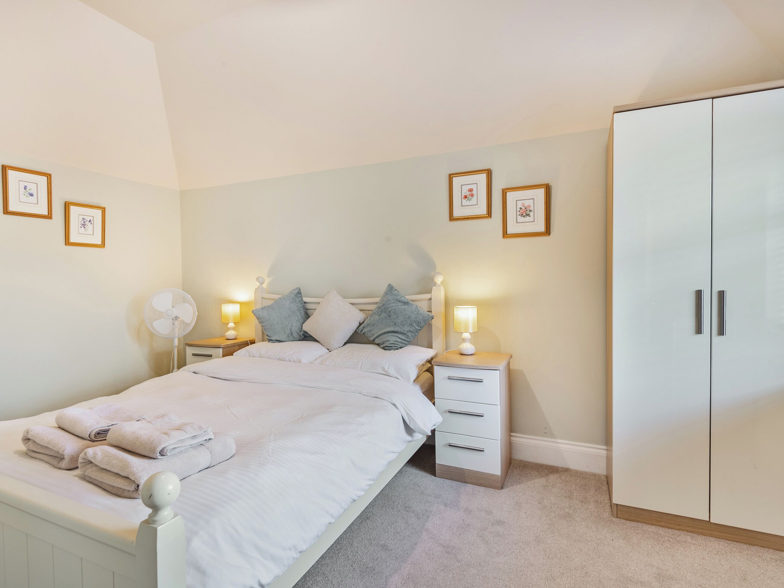 A bedroom with a bed, bedside tables with lamps, a wardrobe, and framed pictures on the wall.