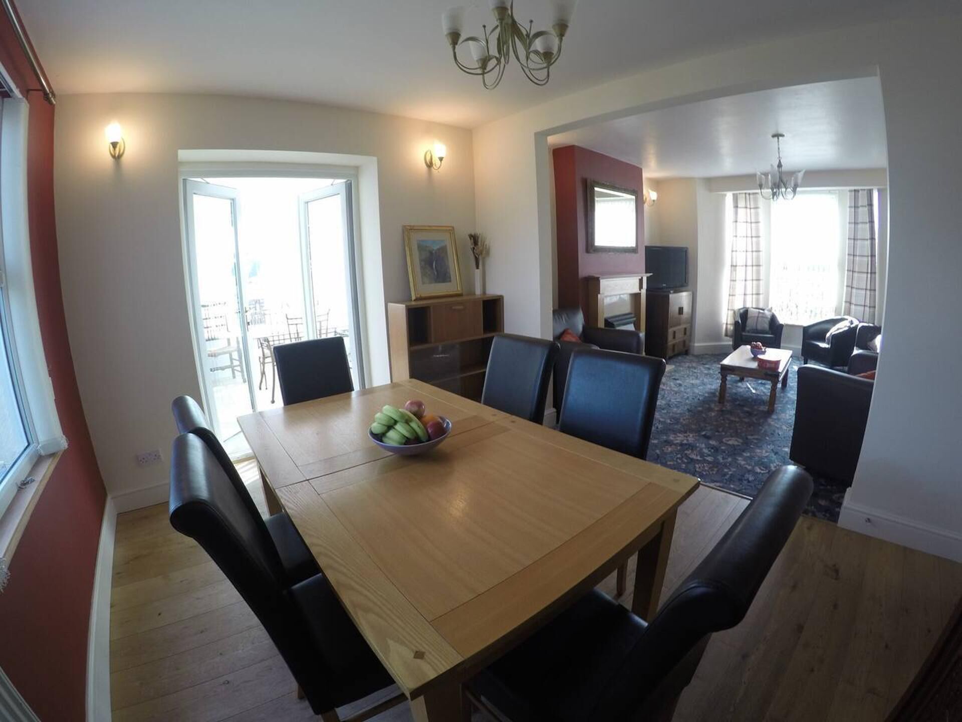 A dining area with a wooden table and black chairs, a fruit bowl on the table, and a television in the background.