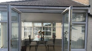 Eine überdachte Terrasse mit einem Glastisch und Stühlen, einem kleinen Blumenvasen und Blick auf die Straße durch die Fenster.