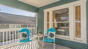 Eine Veranda mit zwei blauen Stühlen, einem kleinen Tisch mit Pflanze und Tasse sowie einem Fenster mit Vorhängen.