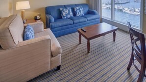Une chambre d’hôtel avec un canapé beige, un canapé bleu agrémenté de coussins à motifs, une table basse en bois et une grande fenêtre donnant sur le port de plaisance.