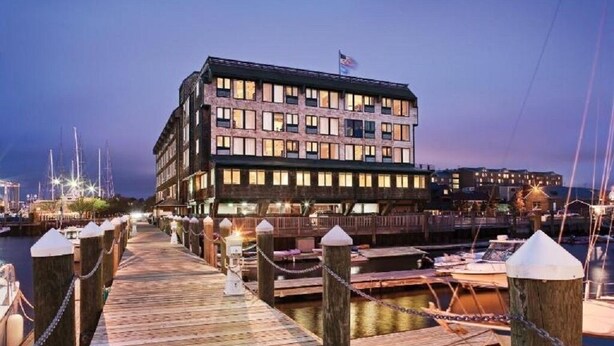 Un hôtel situé au bord de l’eau, doté d’une terrasse en bois menant au quai, d’un drapeau sur le bâtiment et de bateaux en arrière-plan.