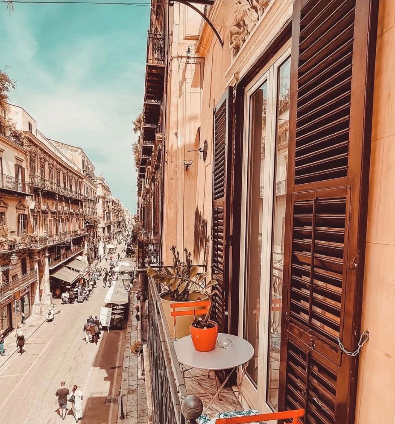 Un balcone con tavolo e sedie, affacciato su una strada con edifici e persone.