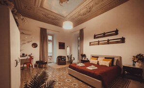 A spacious bedroom with a large bed, a desk, and a chair. There is a window with curtains, a plant, and a decorative ceiling.