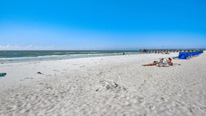 En strand med en brygga i bakgrunden, en sandstrand och människor som kopplar av.