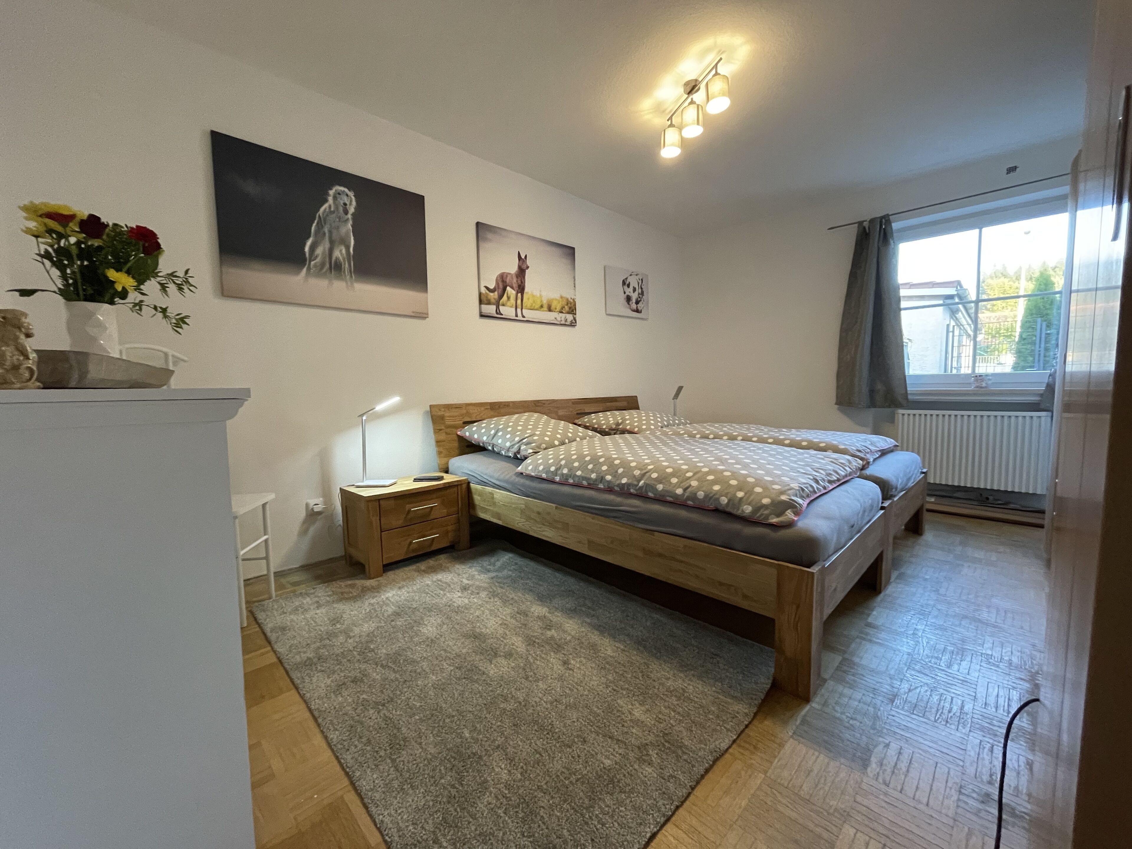 Een slaapkamer met een houten bed, een nachtkastje, een bureau en een raam met gordijnen.