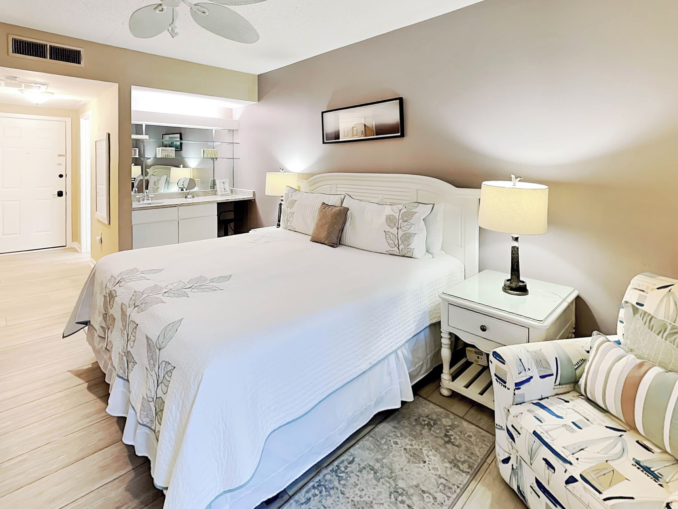A hotel room with a large bed, a bedside table with a lamp, a chair with patterned upholstery, a small desk with a chair, and a bathroom visible through an open door.